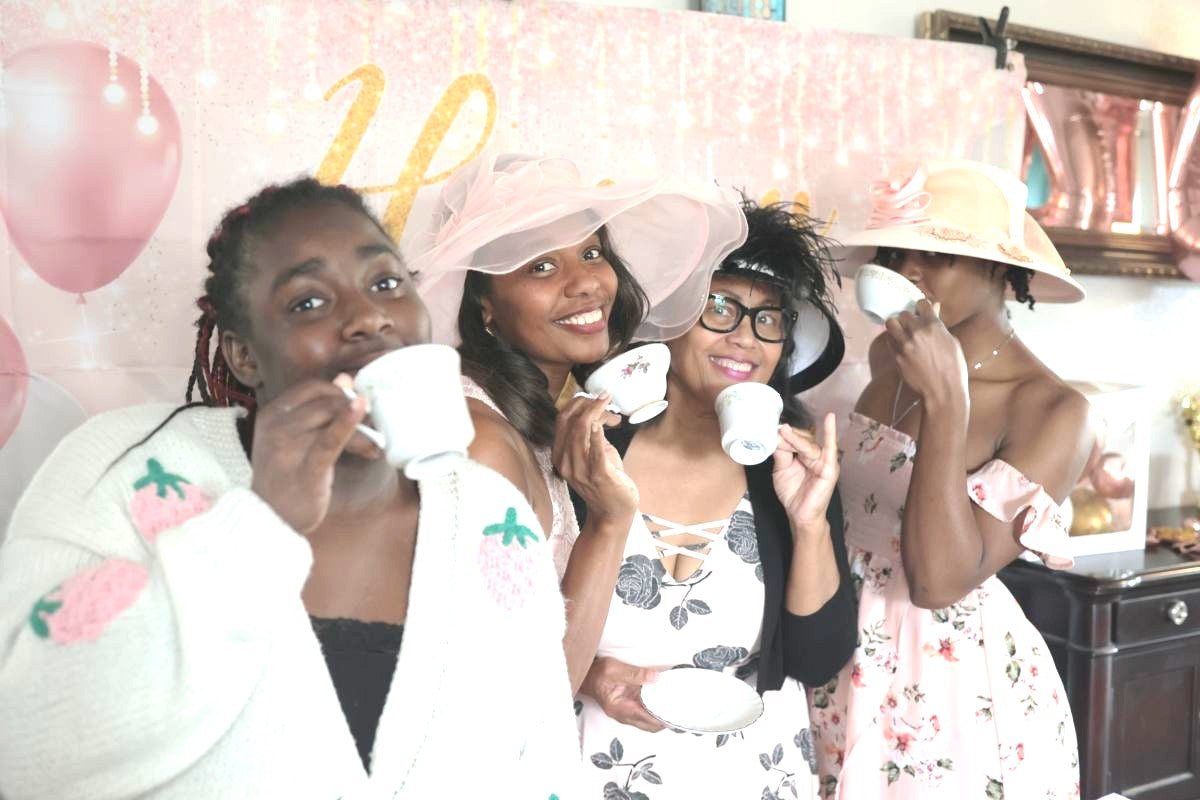 Four women posing together with decorative hats and a pink background