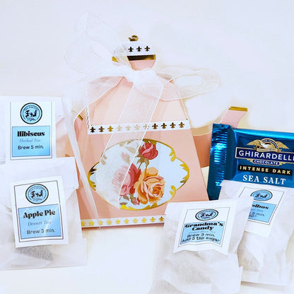 A mini teapot sampler set with four different tea flavors and a chocolate candy, packaged in a pink gift box with a floral design and a white ribbon.