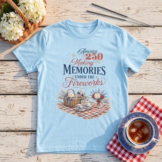 Baby Blue America 250 Making Memories Fourth of July shirt laid flat on a picnic table beside a teacup and flowers