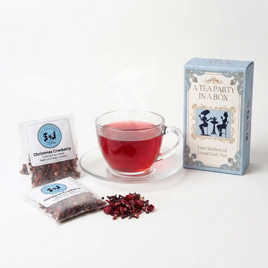 Christmas Cranberry tea brewed in a clear glass teacup beside two labeled loose leaf tea sachets and the A Tea Party In A Box gift box