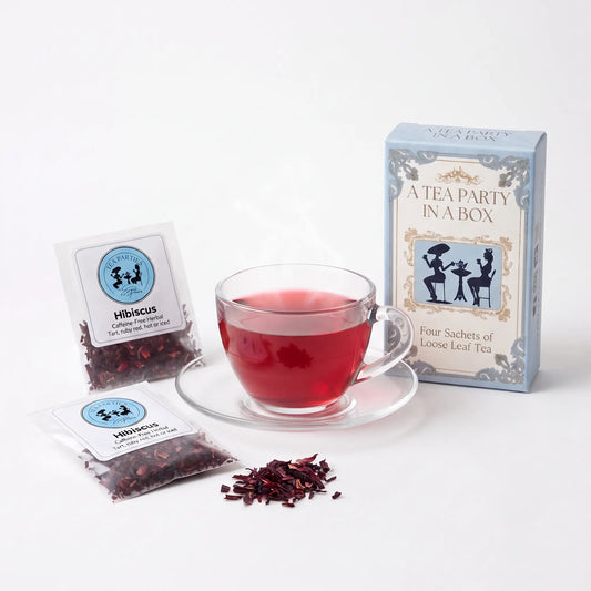 Hibiscus tea brewed in a clear glass teacup beside two labeled loose leaf tea sachets and the A Tea Party In A Box gift box