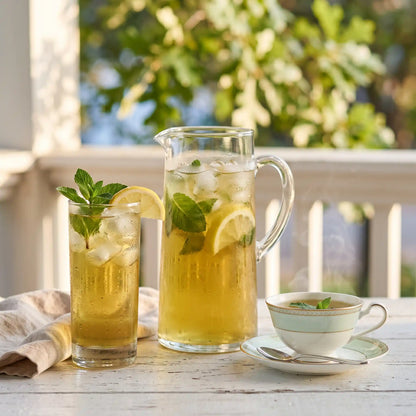 A tall glass of iced mint green tea with mint and lemon beside a pitcher on a sunlit porch table