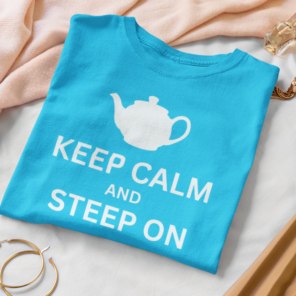 A folded bright blue T-shirt with a white teapot graphic and the words 'KEEP CALM AND STEEP ON' is styled on a white surface with a pink scarf, gold hoop earrings, a pink quilted pouch, and a tan pleated skirt.