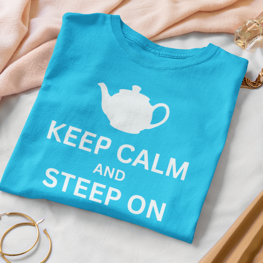 A folded bright blue T-shirt with a white teapot graphic and the words 'KEEP CALM AND STEEP ON' is styled on a white surface with a pink scarf, gold hoop earrings, a pink quilted pouch, and a tan pleated skirt.