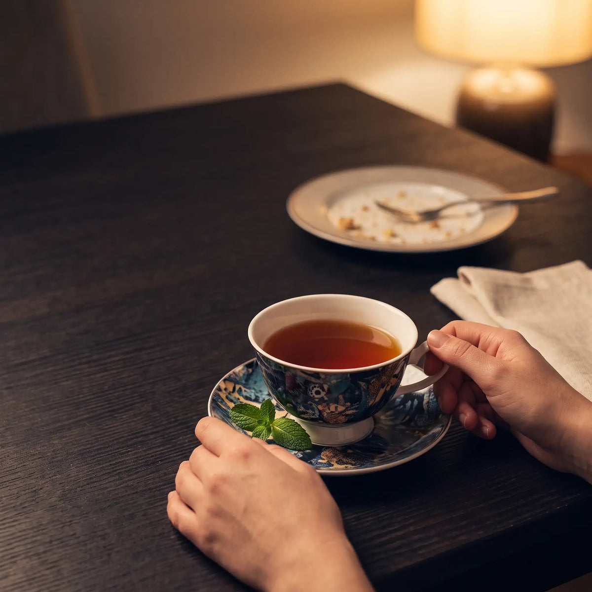 Hands around a white cup of dark tea on a cleared dinner table with a dessert plate pushed aside