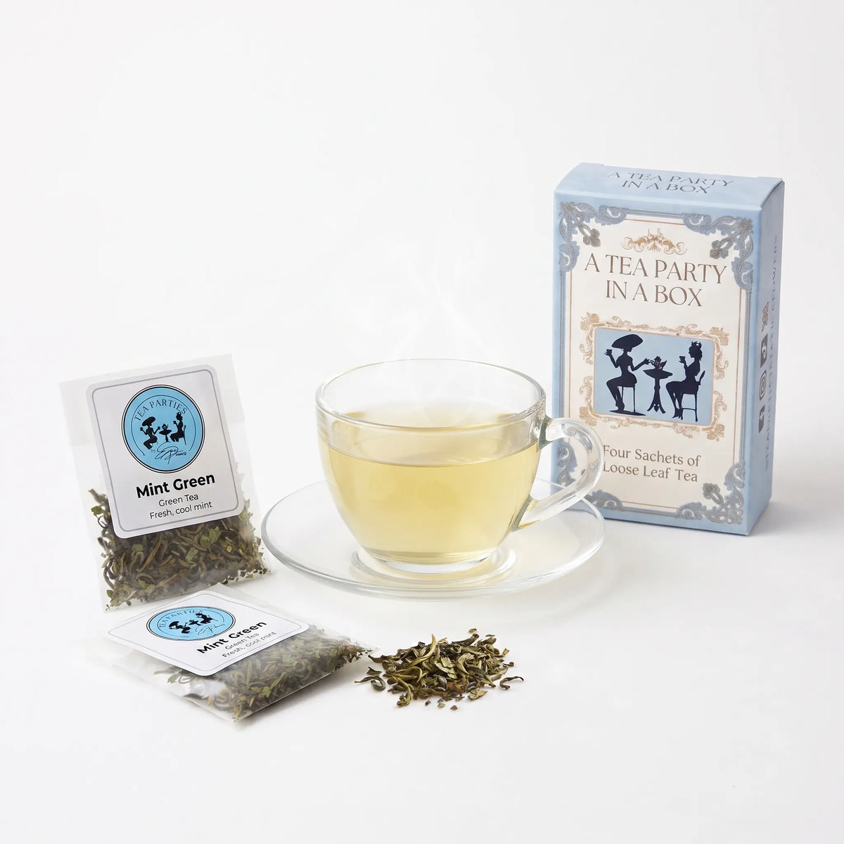 Mint Green tea brewed in a clear glass teacup beside two labeled loose leaf tea sachets and the A Tea Party In A Box gift box