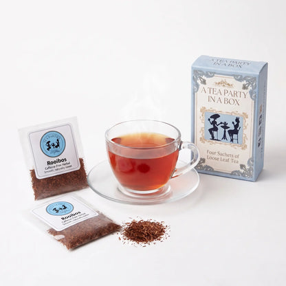 Rooibos tea brewed in a clear glass teacup beside two labeled loose leaf tea sachets and the A Tea Party In A Box gift box