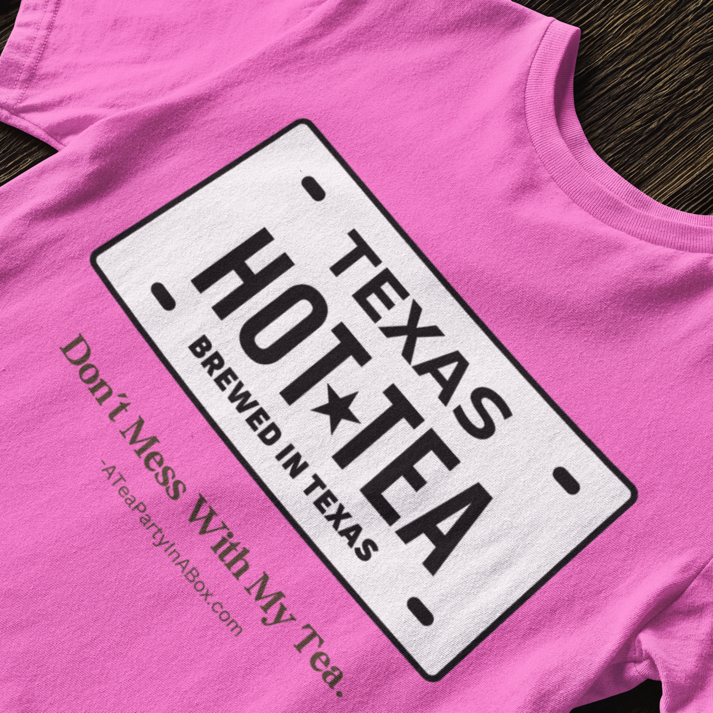 A bright pink t-shirt laid flat on a dark wood background, featuring a white graphic styled like a Texas license plate with the text 'TEXAS HOT TEA BREWED IN TEXAS' and smaller text 'Don't Mess With My Tea. TeaPartyInABox.