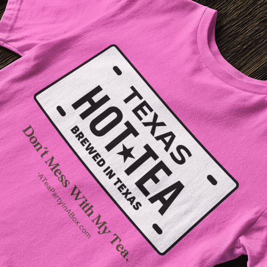 A bright pink t-shirt laid flat on a dark wood background, featuring a white graphic styled like a Texas license plate with the text 'TEXAS HOT TEA BREWED IN TEXAS' and smaller text 'Don't Mess With My Tea. TeaPartyInABox.