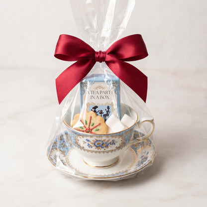 Teacup and saucer gift set wrapped in cellophane with a red bow, a floral cup holding a tea box on a marble counter