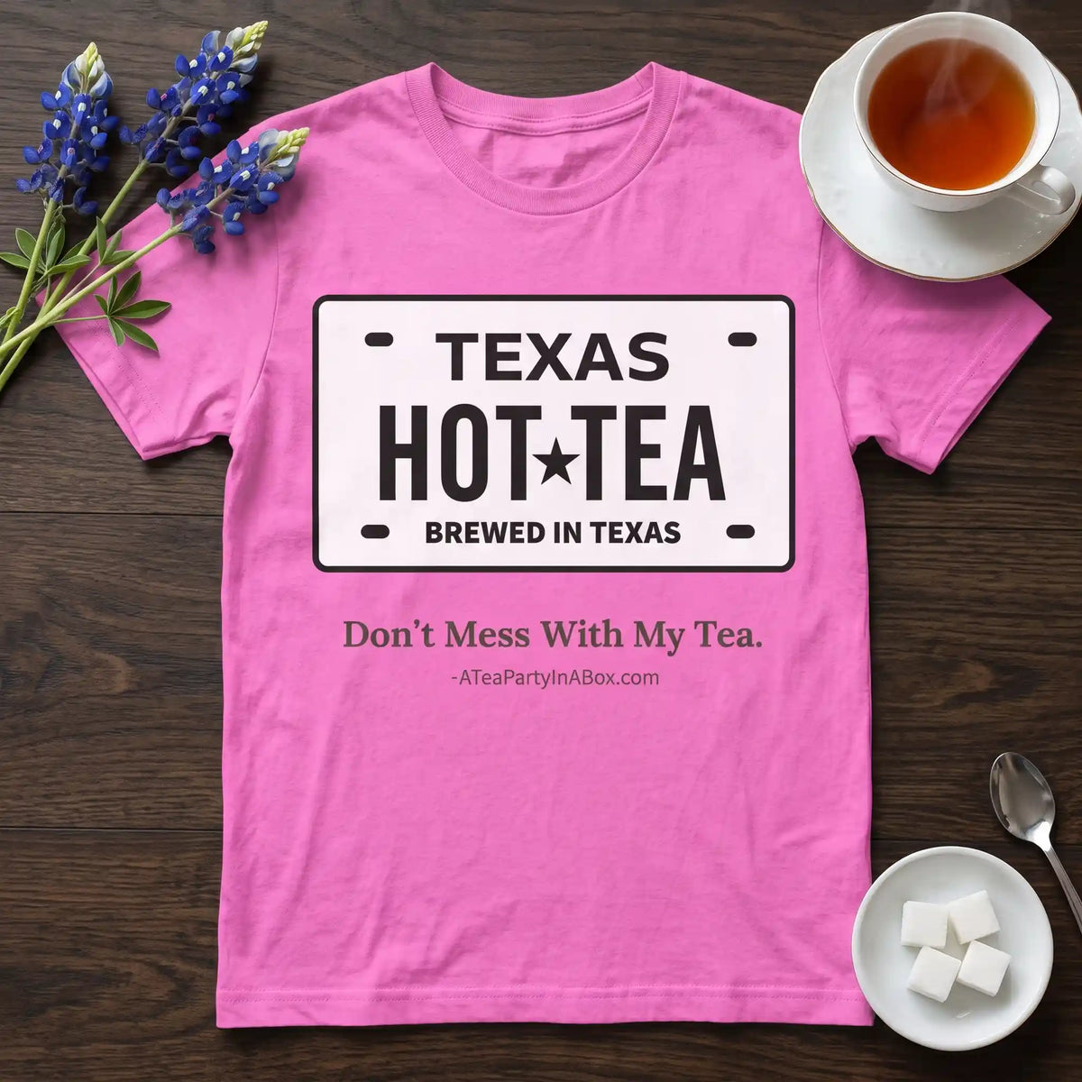 Pink Texas tea shirt with a Hot Tea, Brewed in Texas license plate print on a wooden table beside a cup of hot tea